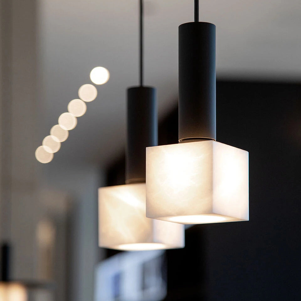 A pair of Clérachic Alabaster Pendant Lights hanging in a dim interior, the stone cubes glowing warm over dark cabinetry.