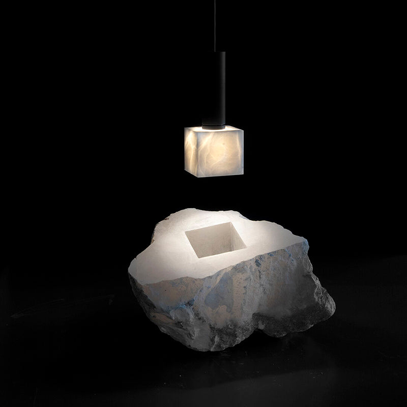 The Clérachic Alabaster Pendant Light lit above the raw quarried block its cube shade was cut from, shown against black.