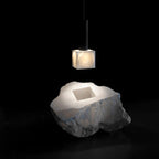The Clérachic Alabaster Pendant Light lit above the raw quarried block its cube shade was cut from, shown against black.