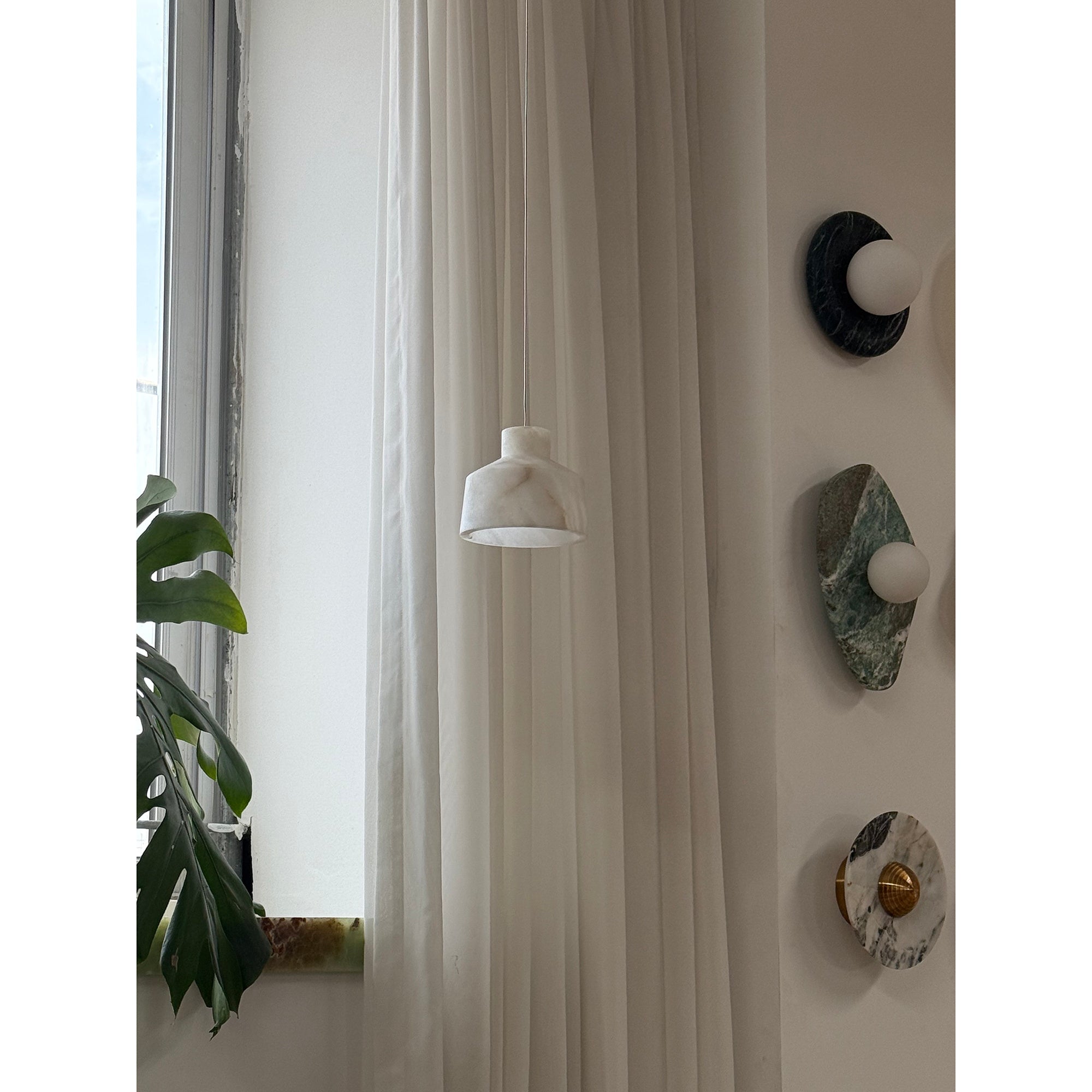 Clérachic Alabaster Pendant Light in white stone hanging beside a curtained window, a monstera leaf in the foreground.
