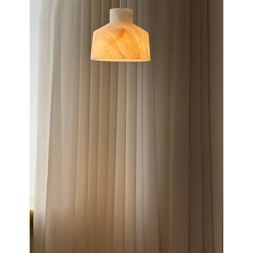 Clérachic Alabaster Pendant Light switched on, warm light travelling through the stone and washing the pleated curtain behind.