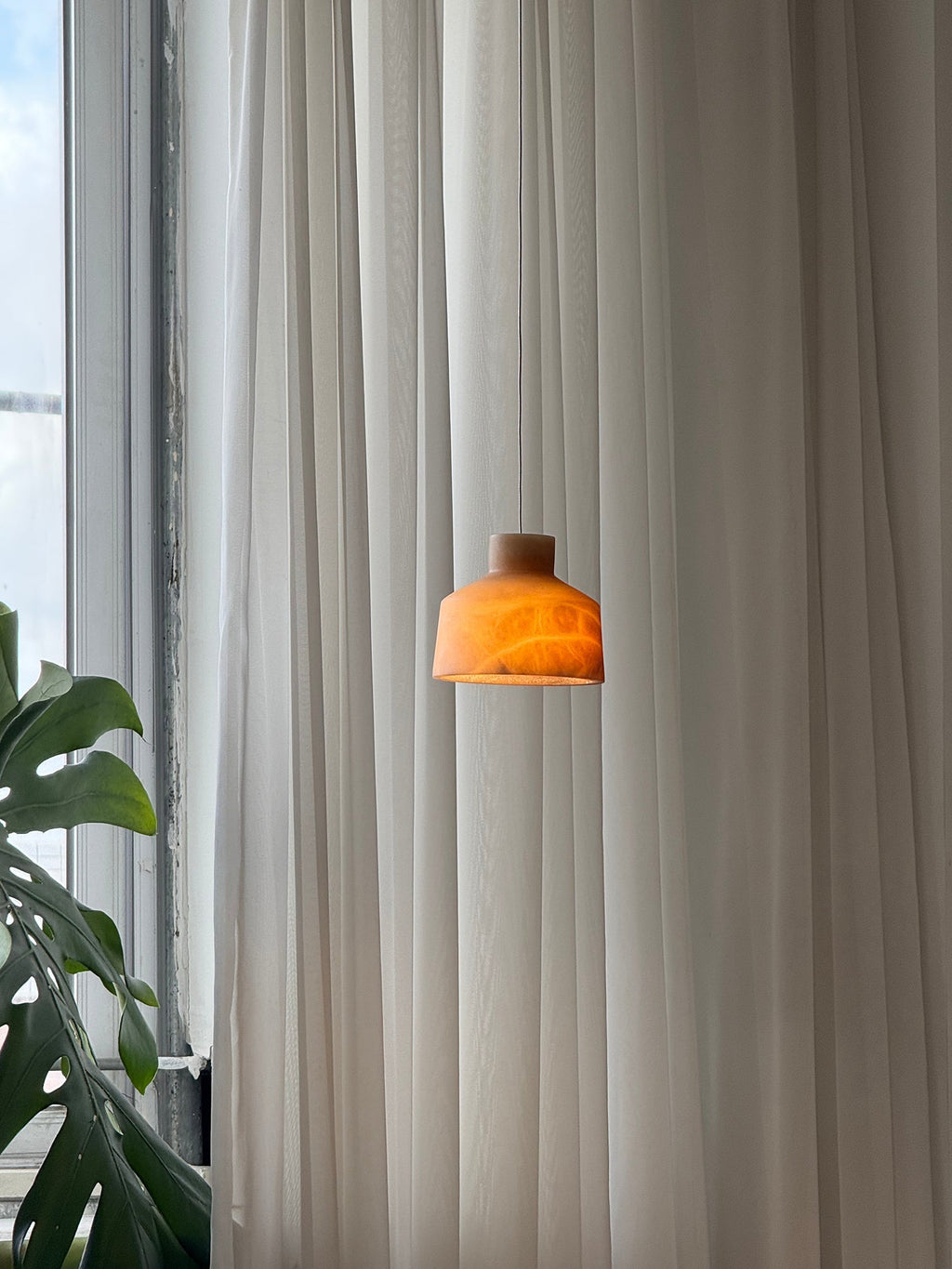 The brown stone Clérachic Alabaster Pendant Light lit at dusk, the shade glowing amber beside a tall window and a monstera.