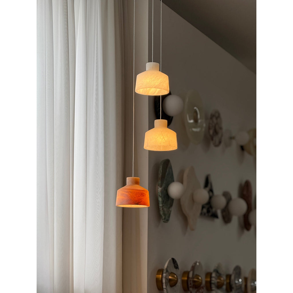 Three-head cluster of the Clérachic Alabaster Pendant Light lit, white, cream and amber shades staggered above a stone-tile wall.