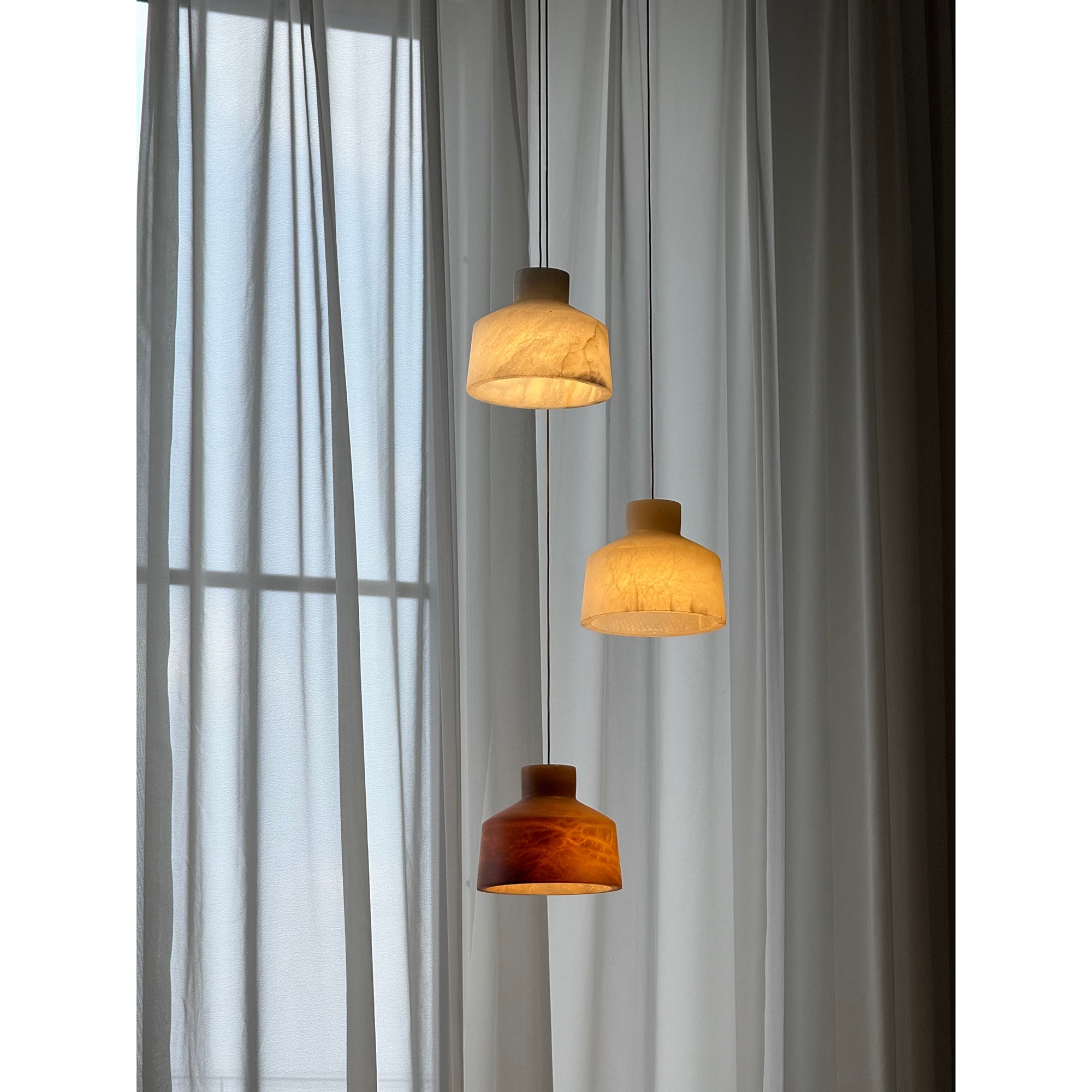 Clérachic Alabaster Pendant Light cluster lit against grey drapes, the lowest shade glowing deep amber below two paler ones.