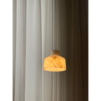 Clérachic Alabaster Pendant Light lit in a dim room, the stone shade glowing gold while the curtain behind falls into shadow.