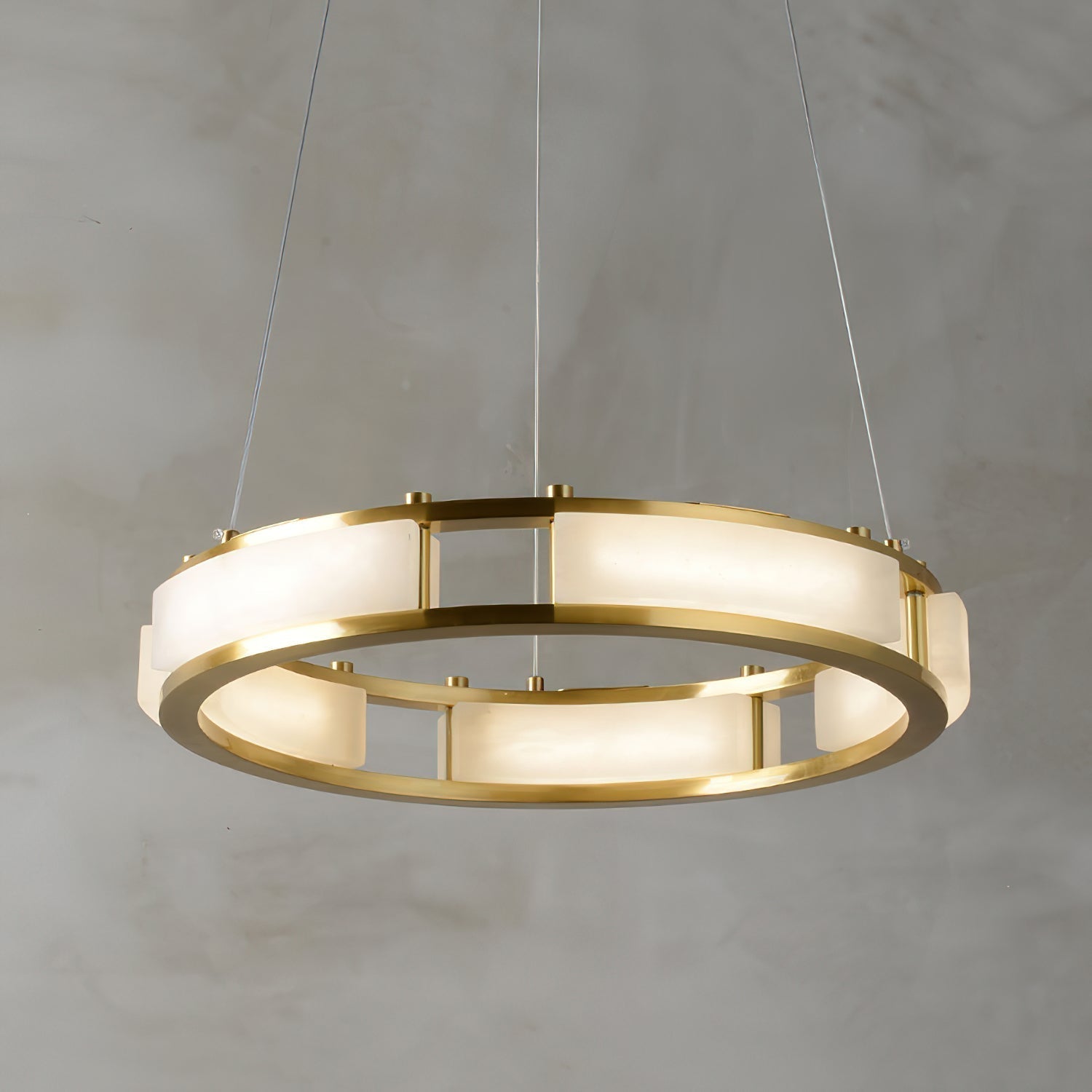 Clérachic Alabaster Ring Chandelier lit against a grey plaster wall, curved stone panels glowing warm between two slim brass rails.