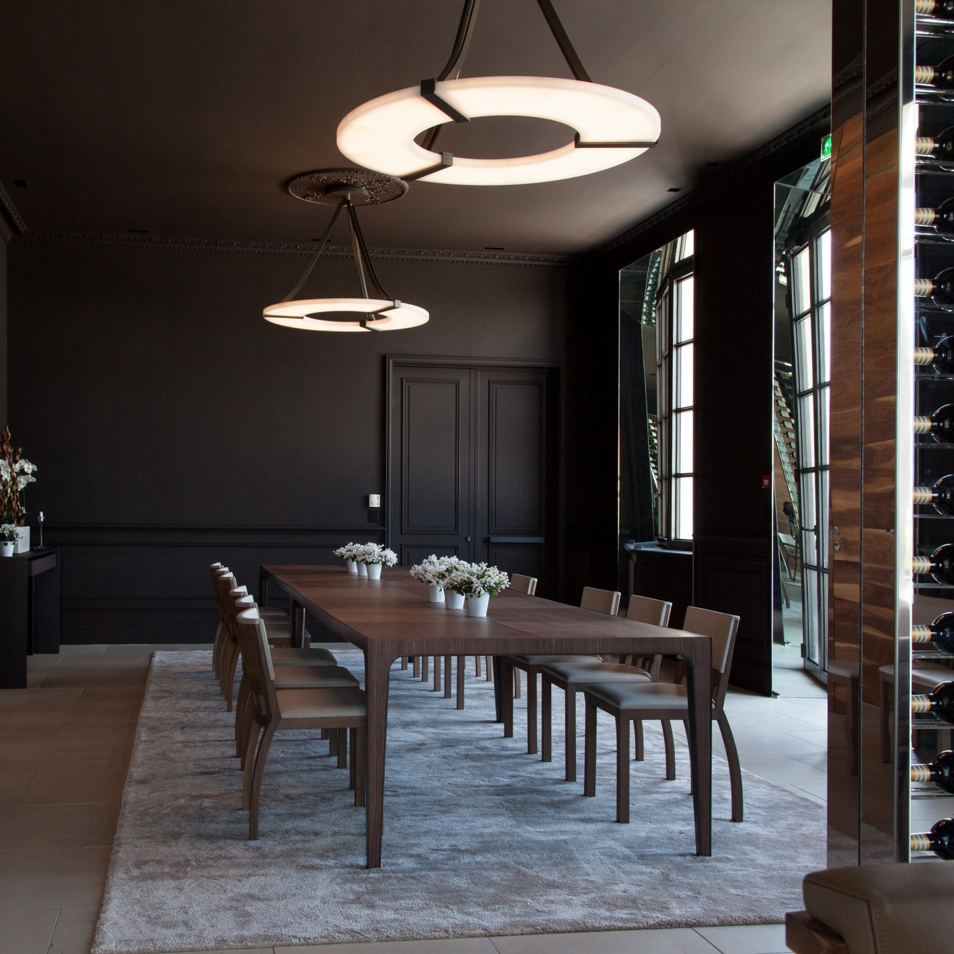 Two Clérachic Alabaster Ring Chandeliers over a long walnut table in a charcoal dining room lined with a mirrored wine wall.