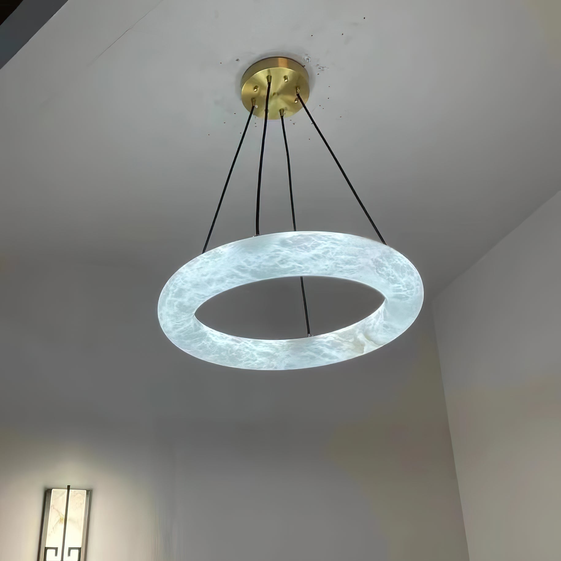 Clérachic Alabaster Ring Chandelier lit from within, the stone glowing crisp white in a hallway with a wall sconce below.