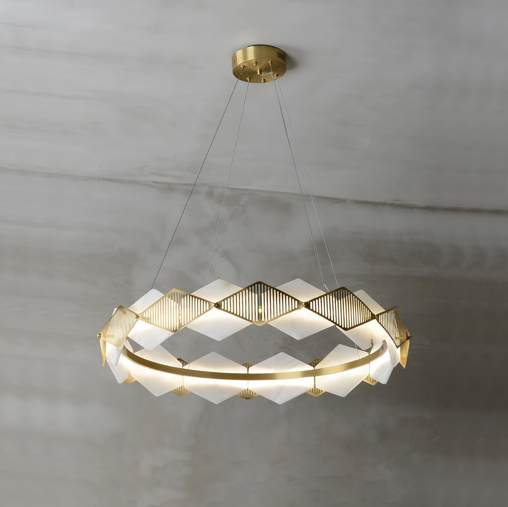 Clérachic Alabaster Ring Chandelier lit on grey plaster, its zig-zag alabaster panels glowing between slotted brass fins.