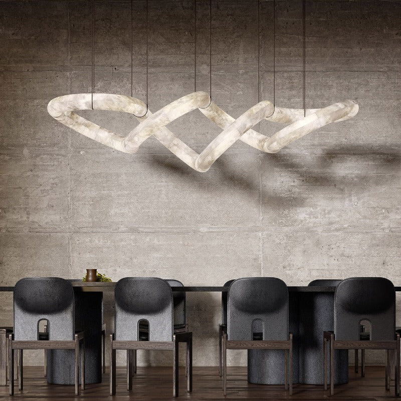 A long Clérachic alabaster ring chandelier stretched above a dark dining table against a raw concrete wall.