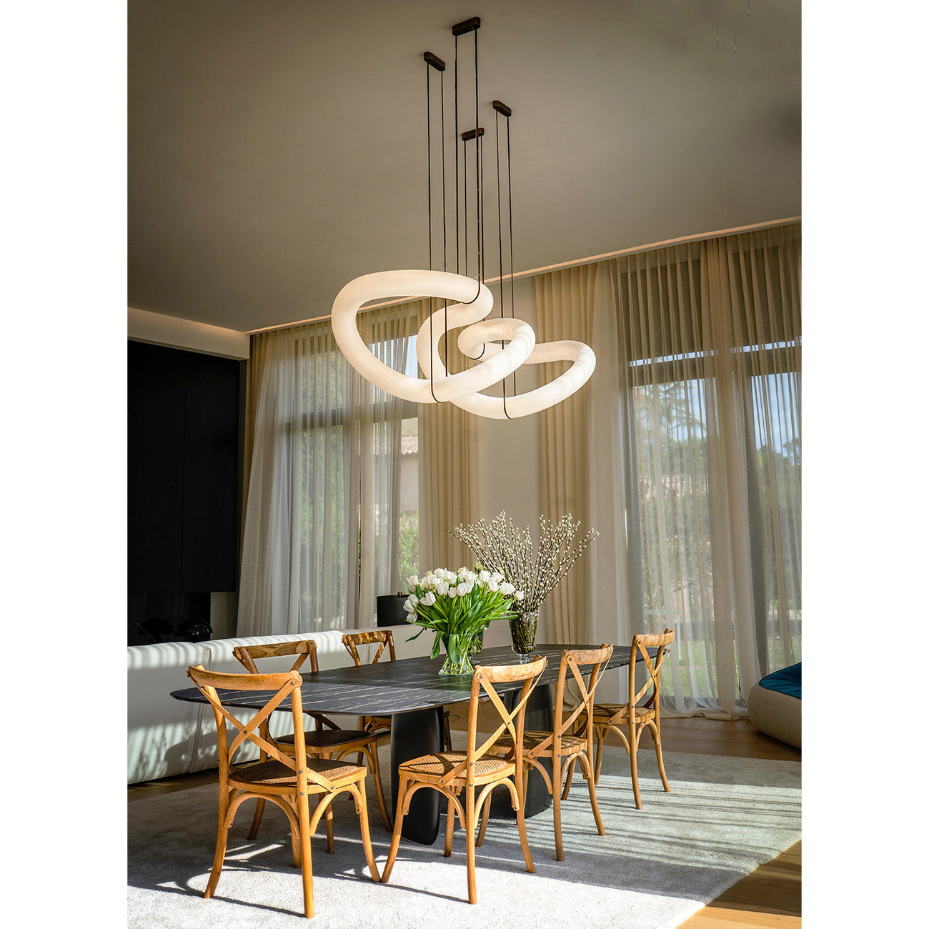 The Clérachic alabaster ring chandelier lit above an oval stone dining table in a sunlit room with sheer curtains.