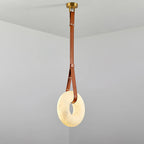 Clérachic Alabaster Ring Pendant Light lit in a bare grey corner, its mid-brown strap looping down from the brass canopy.
