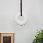 Unlit, the Clérachic Alabaster Ring Pendant Light reads as a pale stone hoop on a dark strap above framed art and a palm.