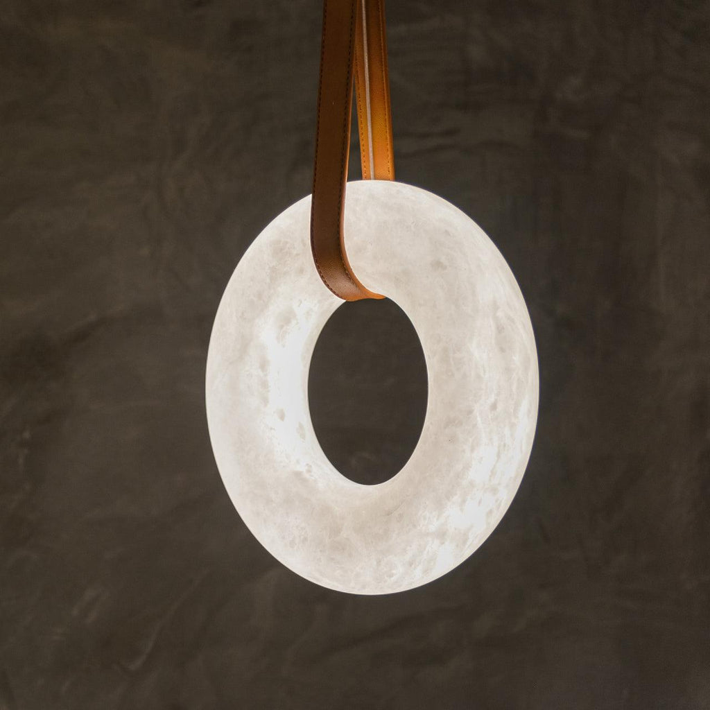 Close view of the Clérachic Alabaster Ring Pendant Light against dark plaster, tan straps looped through the glowing veined stone.