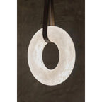 The Clérachic Alabaster Ring Pendant Light glowing against a dark plaster wall, dark leather looped through the stone centre.