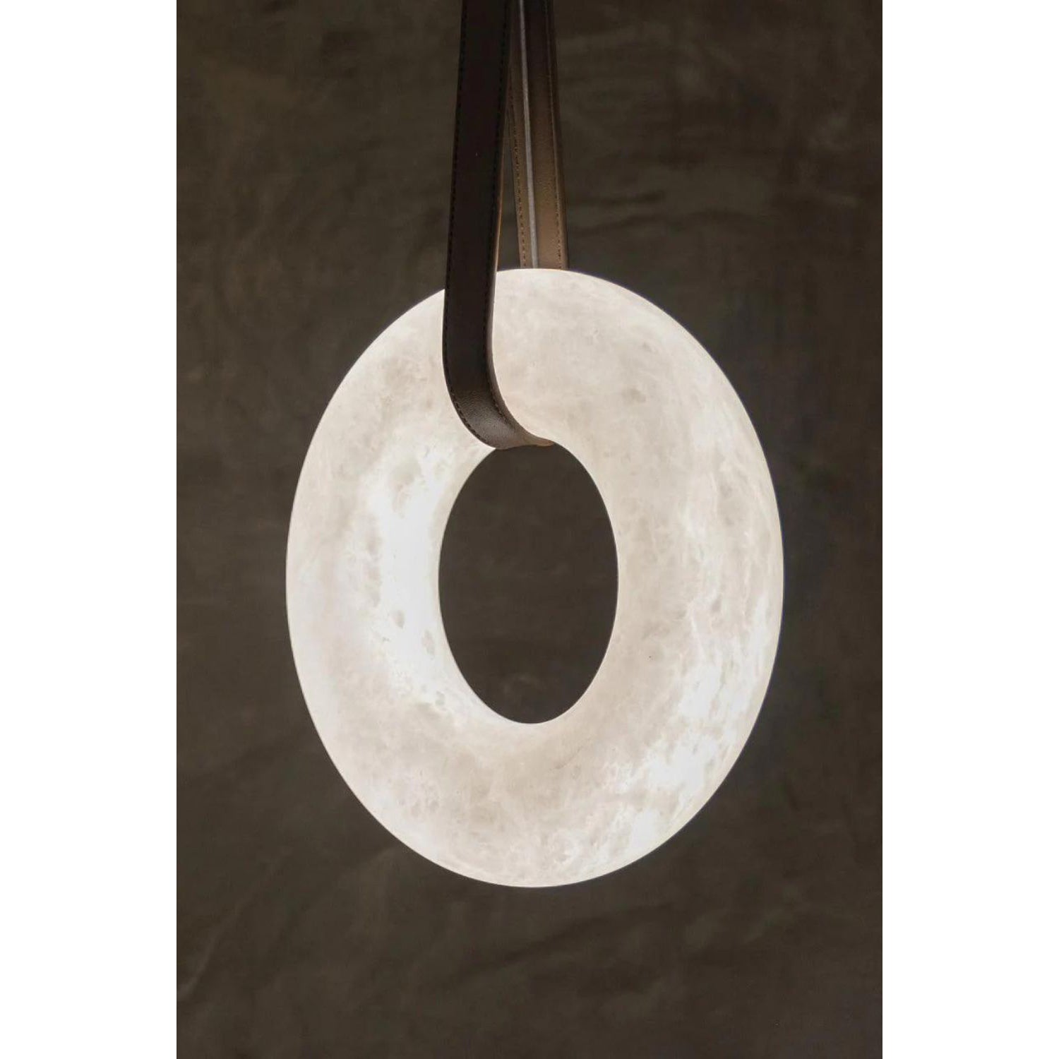 The Clérachic Alabaster Ring Pendant Light glowing against a dark plaster wall, dark leather looped through the stone centre.
