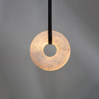 Clérachic Alabaster Ring Pendant Light on a single black strap against a grey backdrop, its ring glowing warm at the centre.
