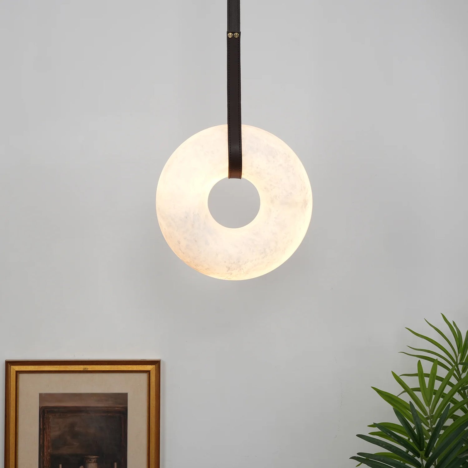 The Clérachic Alabaster Ring Pendant Light glowing beside a gilt-framed print, hung close to the wall on a slim dark strap.