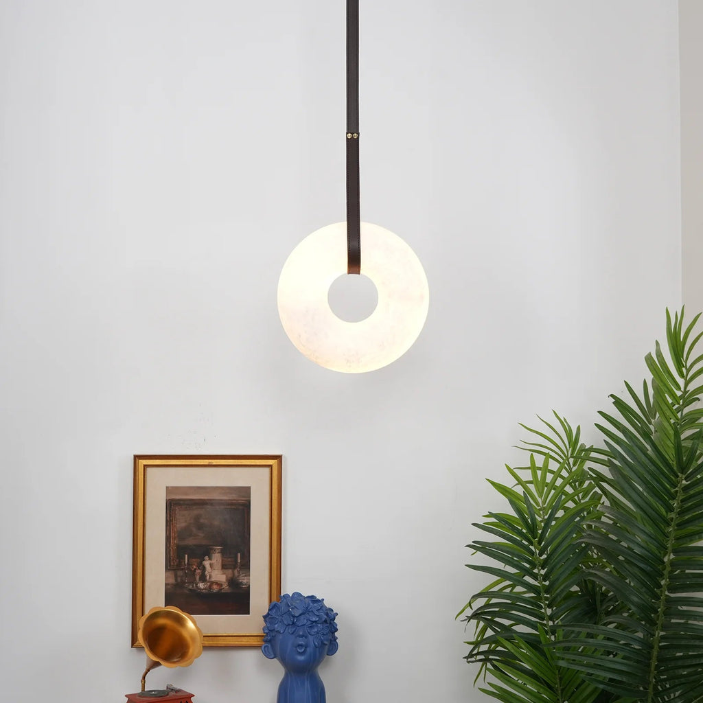 Wide view of a room corner with the Clérachic Alabaster Ring Pendant Light lit above a gilt print and a blue ceramic bust.