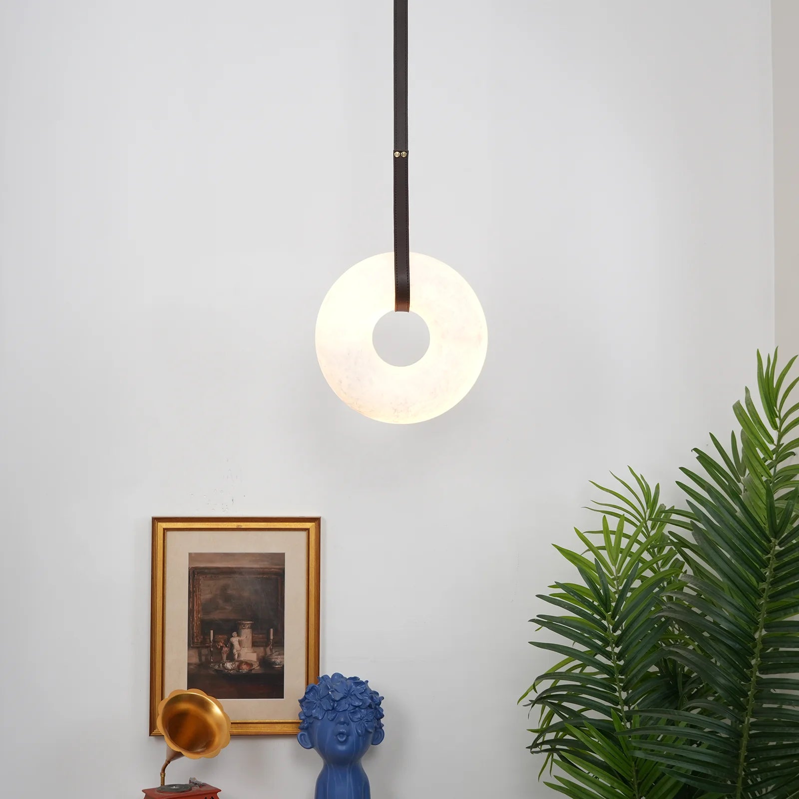 Wide view of a room corner with the Clérachic Alabaster Ring Pendant Light lit above a gilt print and a blue ceramic bust.