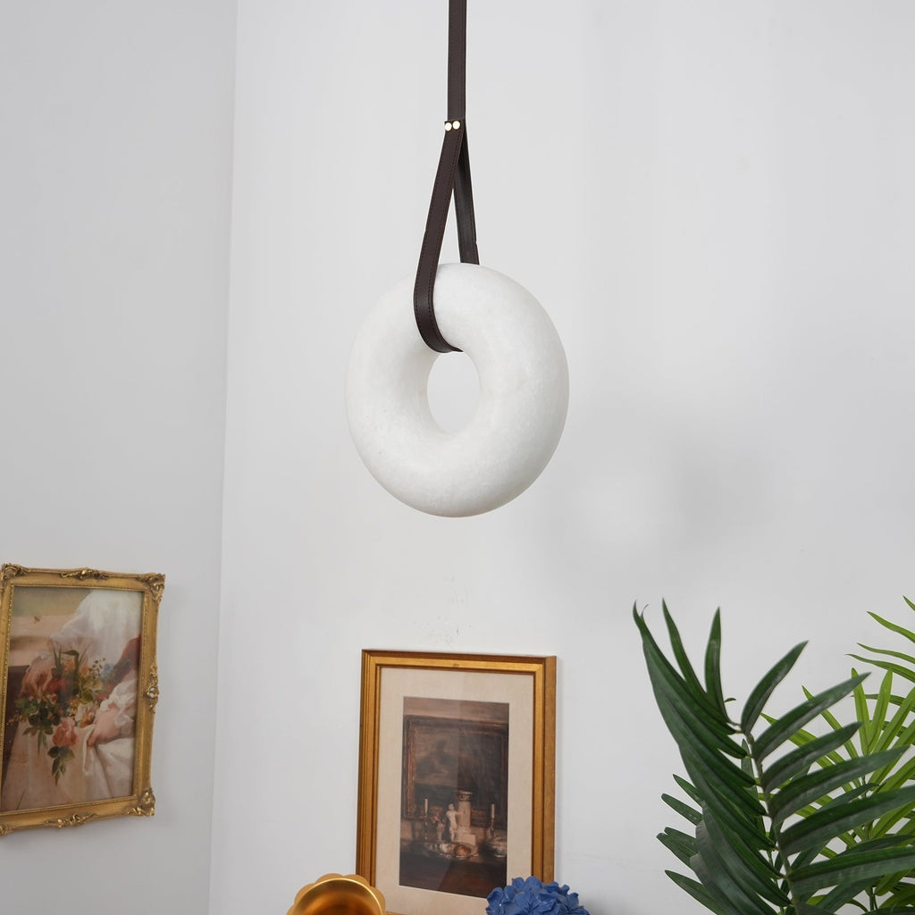 Switched off, the Clérachic Alabaster Ring Pendant Light hangs as plain white stone on a looped strap between two framed prints.