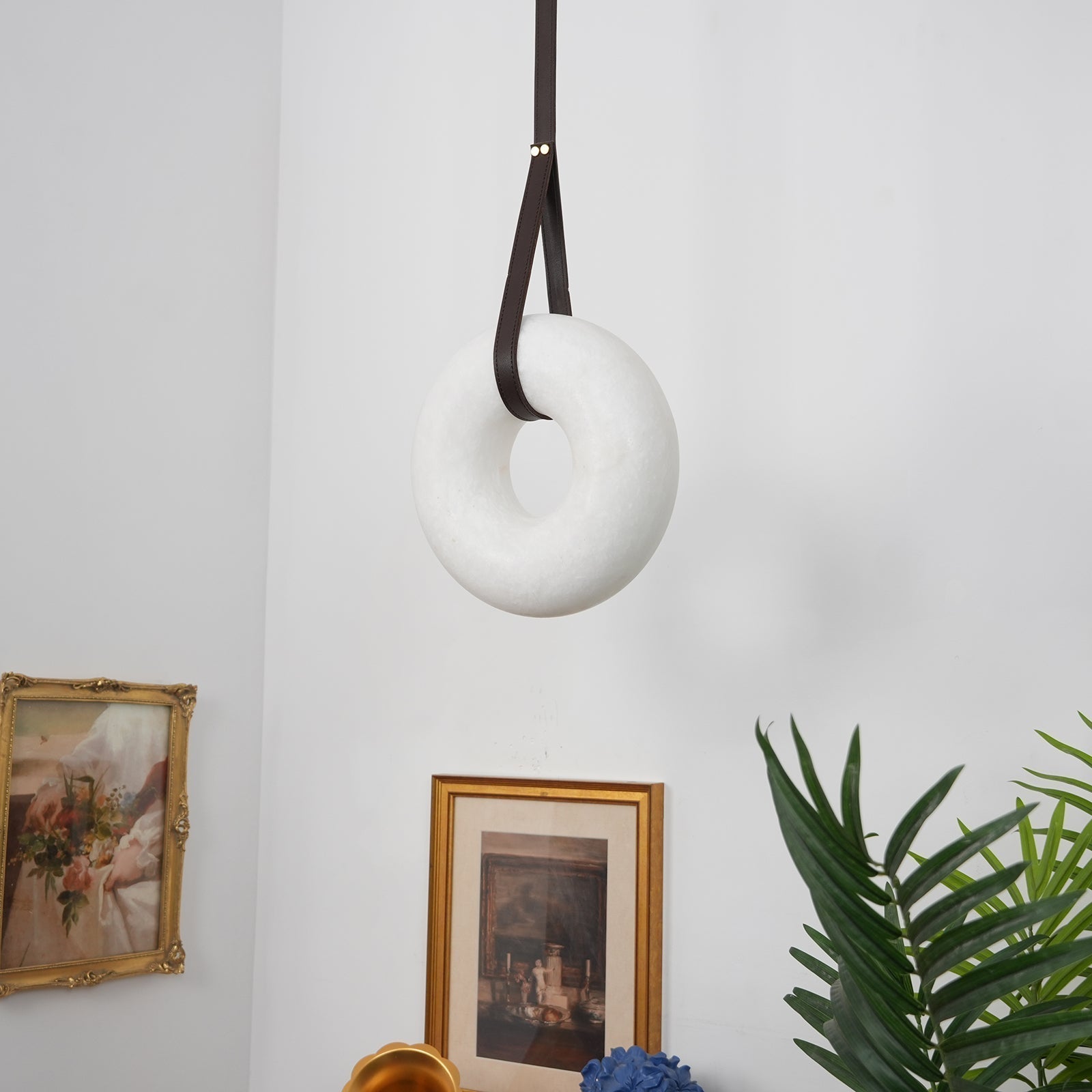 Switched off, the Clérachic Alabaster Ring Pendant Light hangs as plain white stone on a looped strap between two framed prints.