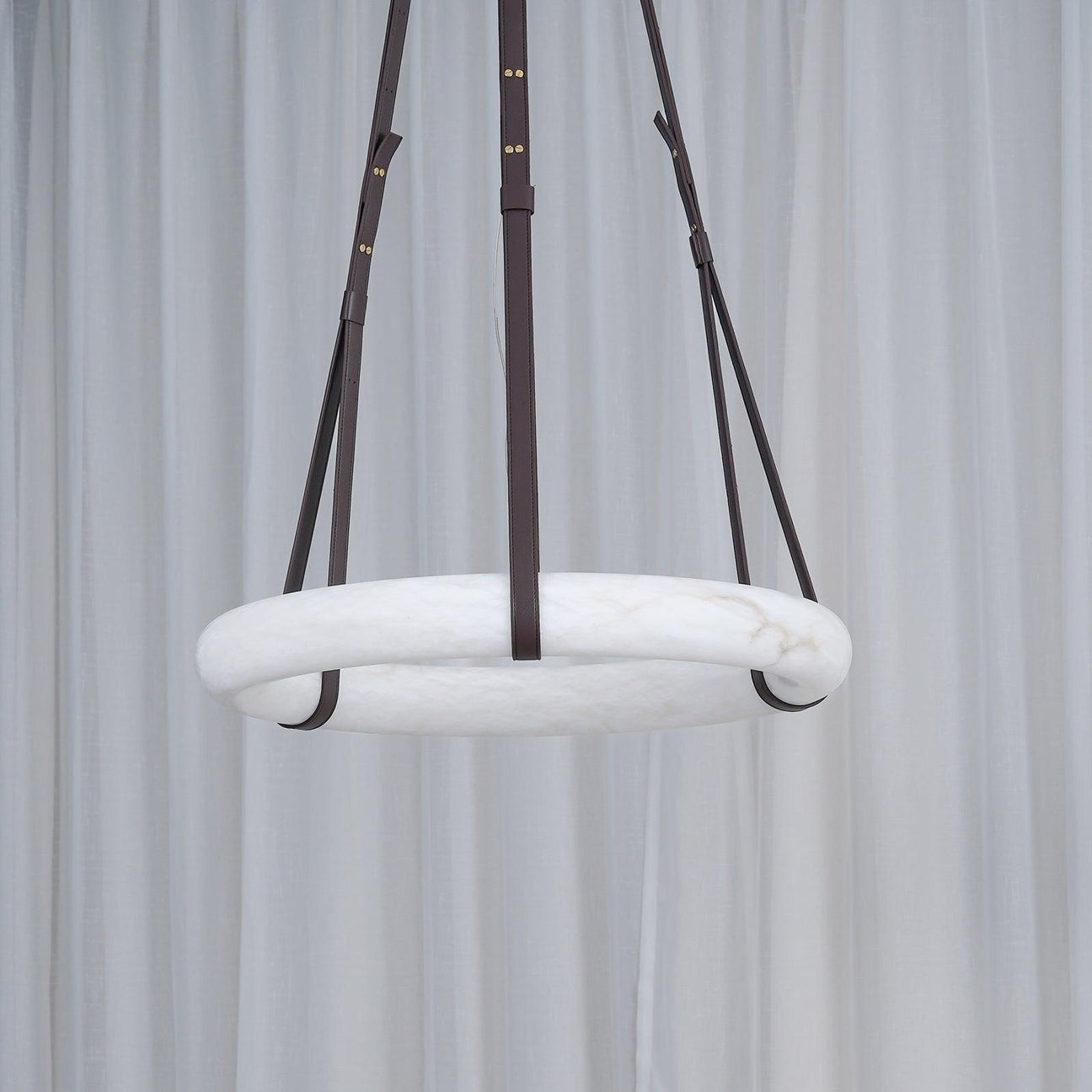 The Clérachic Alabaster Ring Pendant Light unlit in front of a sheer white curtain, reading as a pale stone hoop on dark straps.