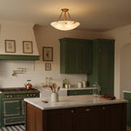 A Clérachic alabaster semi-flush light lit in a kitchen with dark green cabinets, a range cooker and a white marble island.