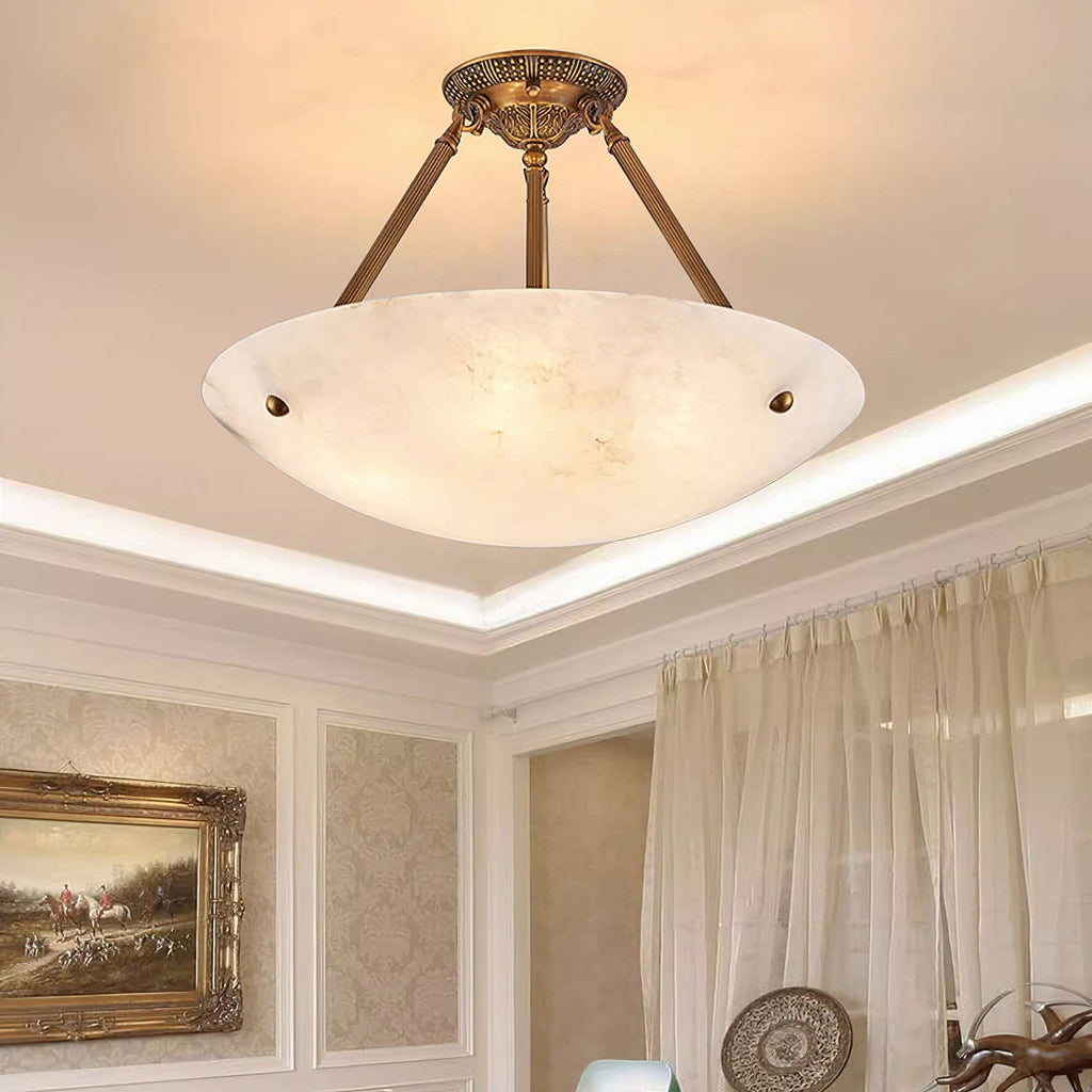 Looking up at a Clérachic alabaster bowl light with brass arms, a lit cove ceiling and sheer curtains beside it.