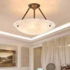 Looking up at a Clérachic alabaster bowl light with brass arms, a lit cove ceiling and sheer curtains beside it.