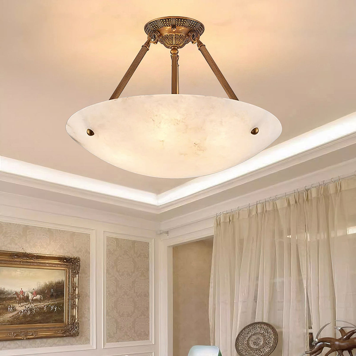 Looking up at a Clérachic alabaster bowl light with brass arms, a lit cove ceiling and sheer curtains beside it.