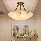 A large Clérachic alabaster bowl light with dark veining hangs in a hall, with a framed horse print and white flowers below.