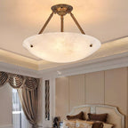 A Clérachic alabaster ceiling light lit above a bedroom with a cream headboard and heavy draped curtains.