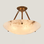 A lit Clérachic alabaster bowl light with antique brass arms and canopy, glowing amber against a pale backdrop.