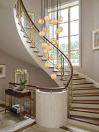 Clérachic alabaster spheres hang by a sweeping staircase with brass balustrade and a tall window, above a console with books and orchids.