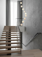 A narrow line of Clérachic alabaster spheres hangs beside an open-tread oak stair with a glass balustrade and grey plaster wall.