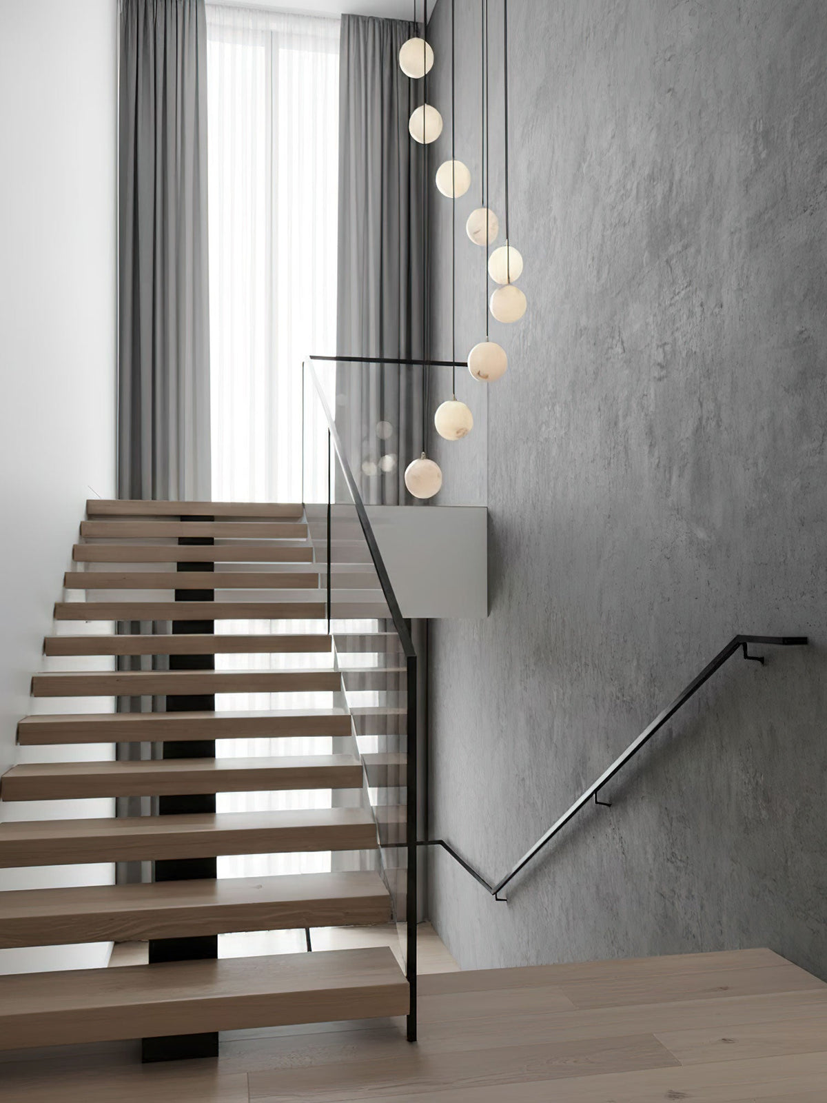 A narrow line of Clérachic alabaster spheres hangs beside an open-tread oak stair with a glass balustrade and grey plaster wall.
