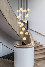 Clérachic alabaster spheres fall in a column beside a curved pale oak stair and a white round wall.