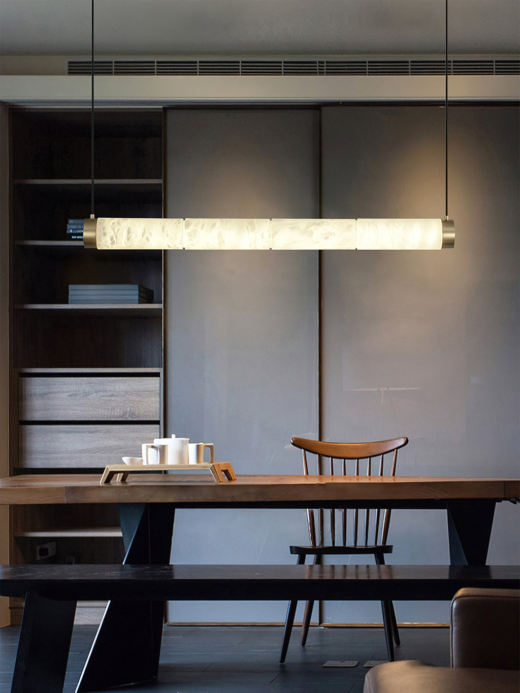 Clérachic linear pendant stretched above a walnut dining table, one continuous bar of warm light in a dark room.