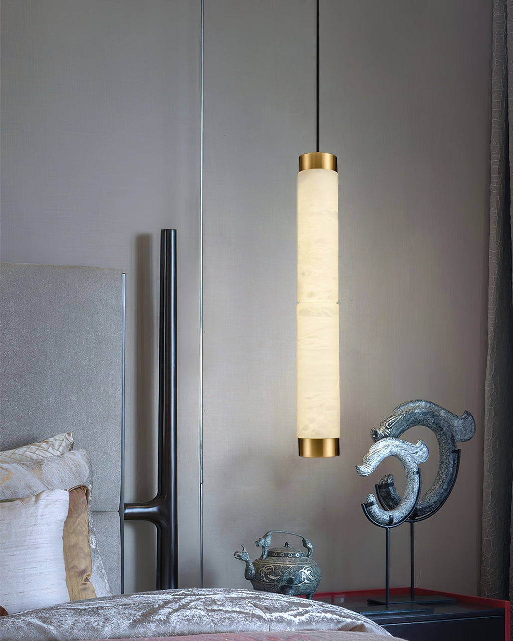 Clérachic pendant dropped beside an upholstered bed, its lit alabaster column framed by a dark bedroom corner.