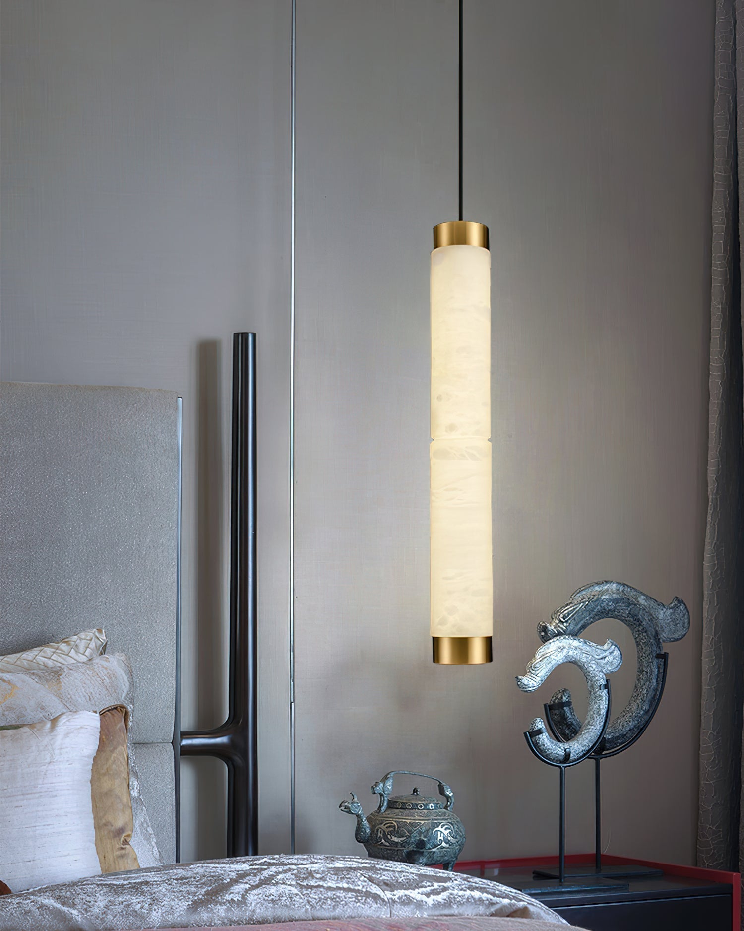 Clérachic pendant dropped beside an upholstered bed, its lit alabaster column framed by a dark bedroom corner.