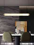 Clérachic strip pendant spanning a dining table, a long bar of lit alabaster against a veined grey stone wall.