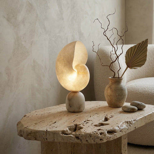 Lit conch-shaped alabaster lamp from Clérachic on a rough-edged travertine side table, beside a stone vase of dried twigs and a pampas-leaf fan.