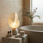 A Clérachic alabaster table lamp glowing on a stone block beside a stone bathtub, with small bottles, a lidded pot and a beaded strand beside it.