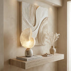 Clérachic conch lamp lit on a floating wooden shelf below a sculpted plaster wall panel, with a book and a vase of dried stems.