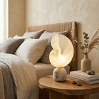 A Clérachic stone and alabaster table lamp on a round wooden bedside table next to a linen-dressed bed with a woven headboard.