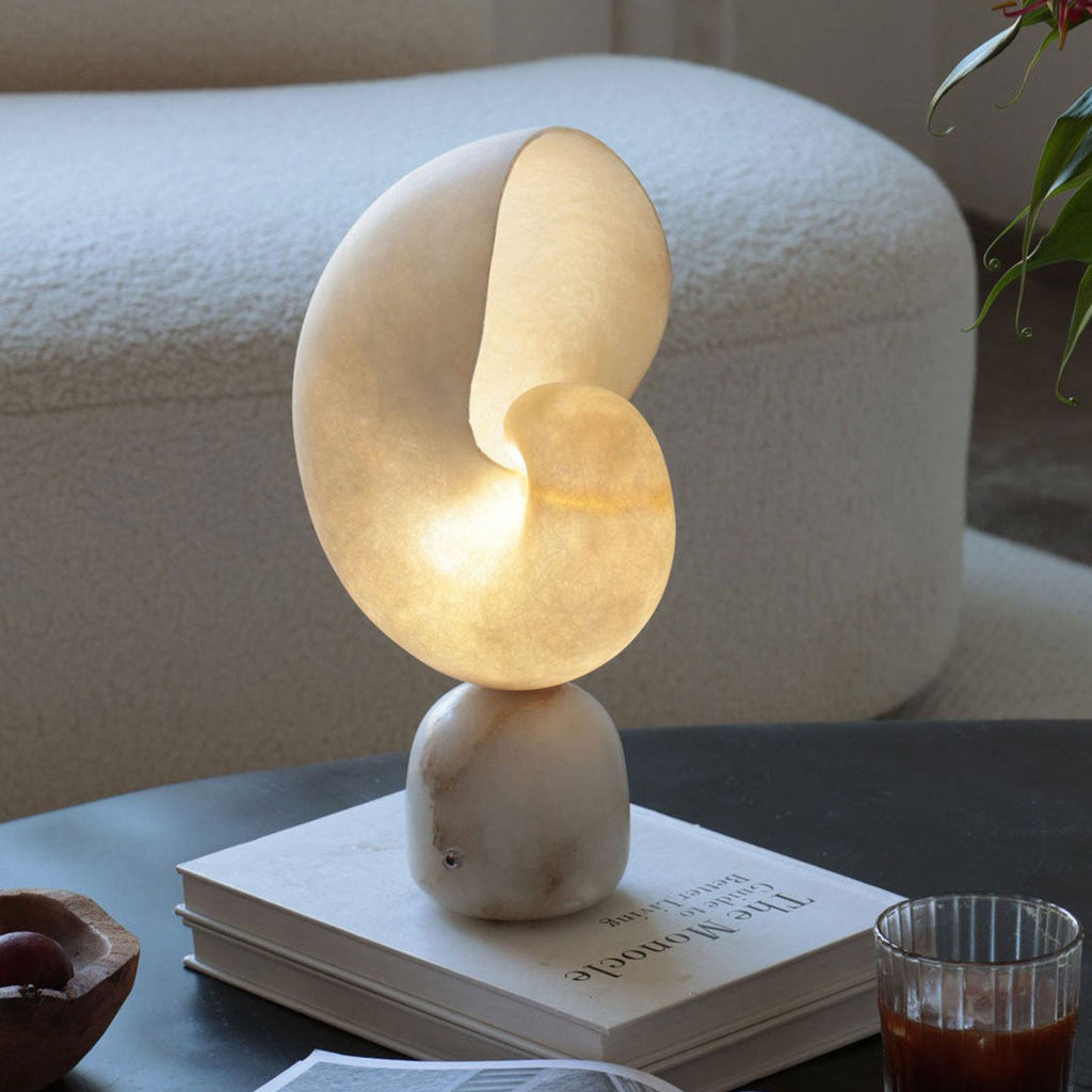 Close view of the glowing alabaster conch shade on this Clérachic table lamp, set on a stack of books on a dark table in front of a boucle sofa.