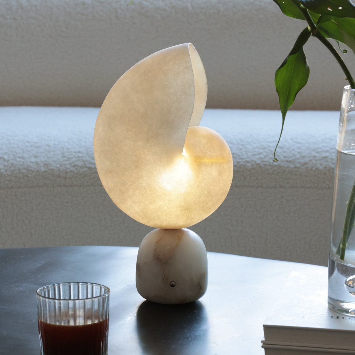 Front view of the Clérachic alabaster lamp with its stone base on a dark table, a glass of tea and a vase of green leaves nearby.