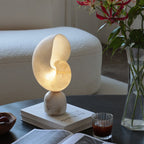 Clérachic conch lamp lit on a stack of books on a dark table, with a vase of red lilies and a glass beside it.