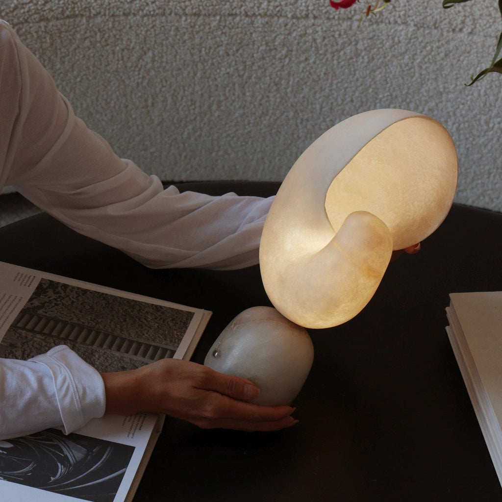 A hand tilting the conch shade of this Clérachic alabaster lamp on its stone base, set on a dark table beside open magazines.